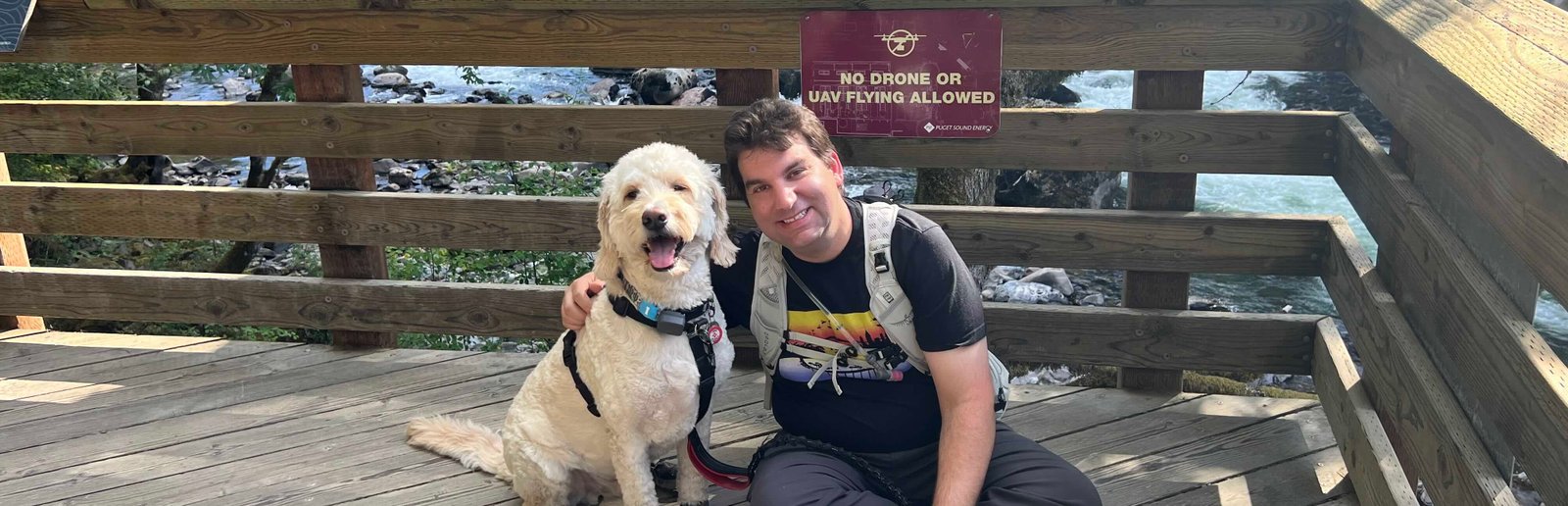 Photo of Ted with his arm around Dexter, a golden mountain doodle, with them both sitting on a boardwalk. Behind them, fastened to the railing, is a sign that reads "No Drone or UAV Flying Allowed."